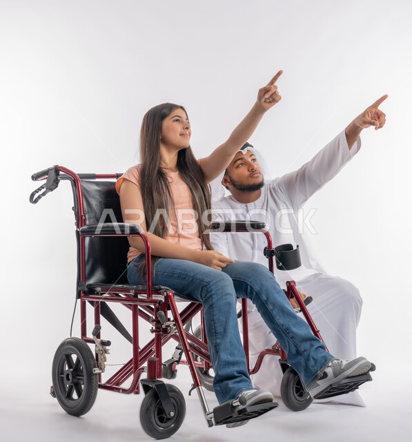 الاشارة إلى شيء ما وتحديد مكان معين, بورتريه لفتاة عربية خليجية اماراتية ترتدي الزي الكاجوال مقعدة تجلس على كرسي متحرك برفقة والدها, مساعدة ودعم المجتمع الاماراتي لذوي الاحتياجات الخاصة, خلفية بيضاء