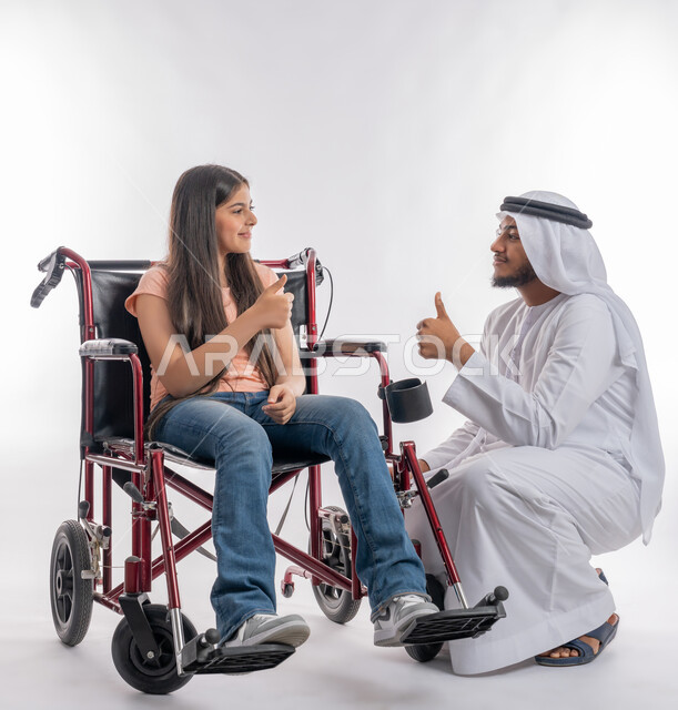 حاجة الأطفال ذوي الهمم إلى الرعاية الصحية الخاصة, تقديم الدعم النفسي للأبناء المعاقين, بورتريه لفتاة عربية خليجية اماراتية مقعدة ترتدي الزي الكاجوال تجلس على كرسي متحرك وترفع اصبعها الابهام بإيماءات الامتياز والموافقة برفقة والدها, خلفية بيضاء