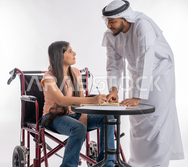 مساعدة أصحاب الهمم على أداء الواجبات المدرسية, مفهوم التعليم المنزلي لذوي الاحتياجات الخاصة, بورتريه لرجل عربي خليجي إماراتي يرتدي الكندورة يقوم بدعم وتشجيع ابنته المقعدة على الدراسة, فتاة إماراتية تجلس على كرسي متحرك, خلفية بيضاء