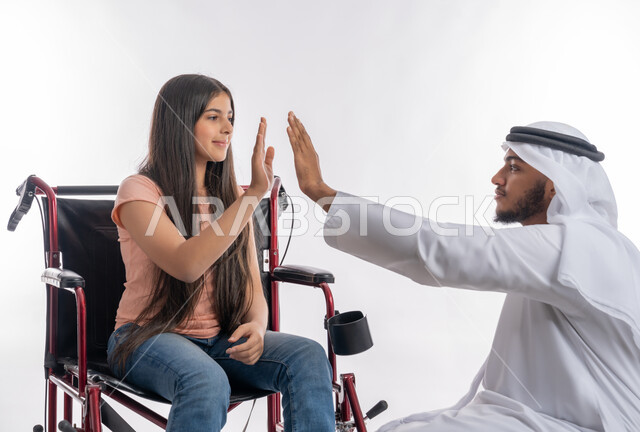 ضرب الكف مقابل الكف للموافقة والإنجاز, الدعم والتشجيع العائلي لذوي الاحتياجات الخاصة, بورتريه لفتاة عربية خليجية اماراتية مقعدة ترتدي الزي الكاجوال تجلس على كرسي متحرك تقوم بالاتفاق مع مع والدها, تمكين المجتمع الاماراتي لأصحاب الهمم, خلفية بيضاء