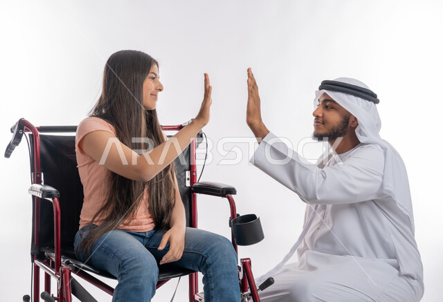 الدعم والتشجيع العائلي لذوي الاحتياجات الخاصة, ضرب الكف مقابل الكف للموافقة والإنجاز, بورتريه لفتاة عربية خليجية اماراتية مقعدة ترتدي الزي الكاجوال تجلس على كرسي متحرك تقوم بالاتفاق مع مع والدها, تمكين المجتمع الاماراتي لأصحاب الهمم, خلفية بيضاء