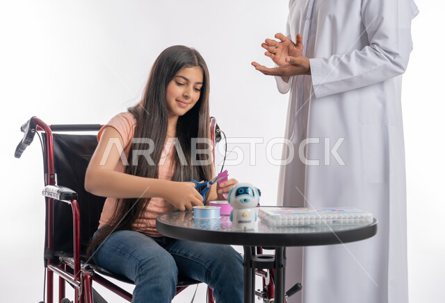 تنمية المهارات والقدرات الحركية لذوي الاحتياجات الخاصة, بورتريه لفتاة عربية خليجية اماراتية مقعدة ترتدي الزي الكاجوال تجلس على كرسي متحرك وتقوم بقص الأوراق الملونة, مفهوم التشجيع والدعم الأسري لذوي الهمم, خلفية بيضاء