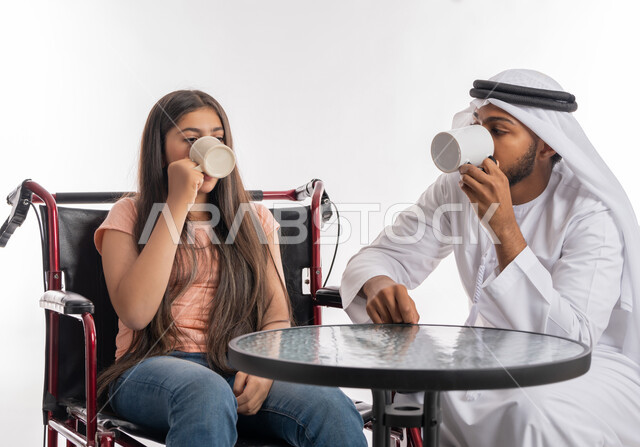 الاستمتاع بتناول المشروبات الساخنة في إحدى المقاهي الخليجية, أجواء أسرية مليئة بالعطف والحب مع أصحاب الهمم, بورتريه لفتاة عربية خليجية اماراتية مقعدة ترتدي الزي الكاجوال تجلس على كرسي متحرك برفقة والدها, مفهوم الاهتمام بذوي الاحتياجات الخاصة, خلفية بيضاء