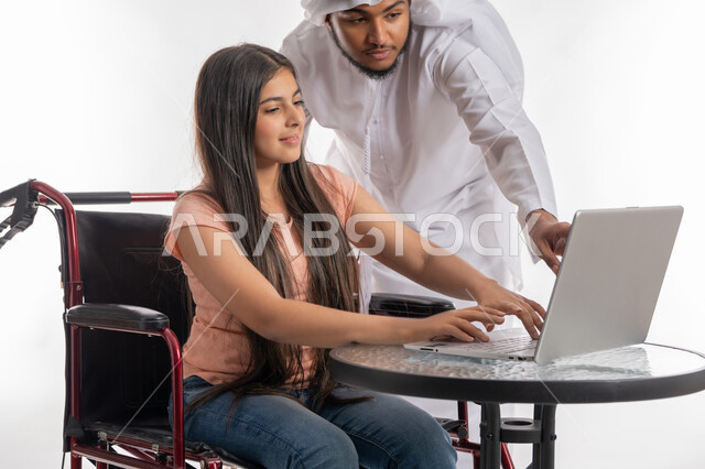 التعليم عبر الأونلاين, قضاء وقت ممتع مع ذوي الاحتياجات الخاصة, بورتريه لفتاة عربية خليجية اماراتية مقعدة ترتدي الزي الكاجوال تجلس على كرسي متحرك تقوم باستخدام الحاسب المحمول برفقة والدها, دمج التقنية والتكنولوجيا مع التعلم, تنمية مهارات ذوي الهمم, خلفية بيضاء
