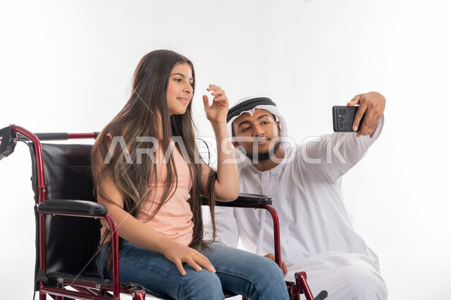 دعم المجتمع الاماراتي لذوي الاحتياجات الخاصة, شعور المعاقين بالأمان والحنان في بيئتهم, بورتريه من الجانب لفتاة عربية خليجية إماراتية ترتدي الزي الكاجوال تقوم بالتقاط صور سيلفي تذكارية برفقة والدها عبر الهاتف المحمول, خلفية بيضاء