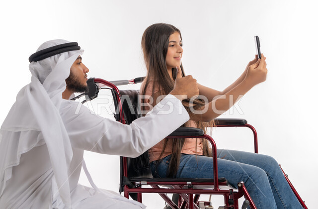 شعور المعاقين بالأمان والحنان في بيئتهم, بورتريه من الجانب لفتاة عربية خليجية إماراتية ترتدي الزي الكاجوال تقوم بالتقاط صور سيلفي تذكارية برفقة والدها عبر الهاتف المحمول, تمكن ودعم المجتمع الاماراتي لذوي الاحتياجات الخاصة, خلفية بيضاء