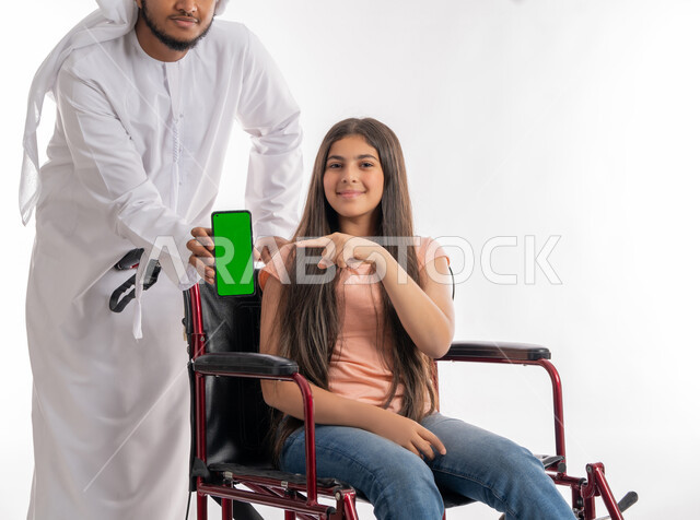 استخدام الأجهزة والتقنيات الحديثة والمتطورة, التواصل مع الأهل والأقارب عبر الجوال, بورتريه لفتاة عربية خليجية إماراتية ترتدي الزي الكاجوال تشير إلى الهاتف المحمول بشاشة خضراء فارغة برفقة والدها, تمكين المجتمع الاماراتي لذوي الاحيتاجات الخاصة, خلفية بيضاء