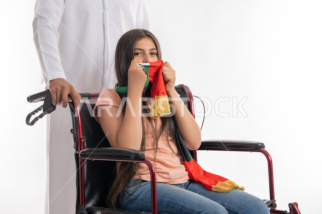 يوم الاستقلال 2 ديسمبر, يوم العلم الاماراتي 3 نوفمير, التباهي بالانتماء للوطن العربي, بورتريه لفتاة عربية خليجية إماراتية ترتدي الزي الكاجوال تجلس على كرسي متحرك وتقوم بتقبيل علم دولة الامارات بفخر واعتزاز برفقة والدها, مساعدة ذوي الهمم على التنقل, خلفية بيضاء