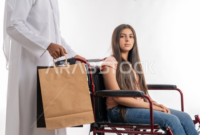 النظر إلى الكاميرا بإيماءات الجدية, قضاء الوقت الممتع بالتسوق برفقة الأبناء, أب عربي خليجي إماراتي يمسك بيده أكياس التسوق ويساعد ابنته على الحركة, بورتريه لفتاة إماراتية ترتدي الزي الكاجوال تجلس على كرسي متحرك, تقديم الدعم والاهتمام لأصحاب الهمم, خلفية بيضاء