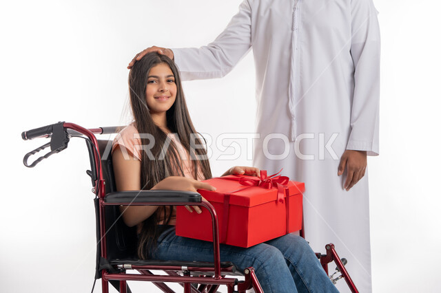 الألفة والمحبة بين أفراد الأسرة, الاهتمام بالأبناء من ذوي الهمم, تقديم الدعم والرعاية الأسرية لأصحاب الإعاقة الحركية, بورتريه لفتاة عربية خليجية إماراتية مقعدة ترتدي الزي الكاجوال تجلس على كرسي متحرك تمسك بيدها علبة هدية حمراء برفقة والدها, خلفية بيضاء