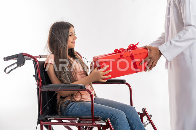 الاهتمام بالأبناء من ذوي الاحتياجات الخاصة, تقديم الدعم والرعاية الأسرية لأصحاب الهمم, بورتريه من الجانب لفتاة عربية خليجية إماراتية مقعدة ترتدي الزي الكاجوال تجلس على كرسي متحرك تمسك بيدها علبة هدية حمراء برفقة والدها, خلفية بيضاء