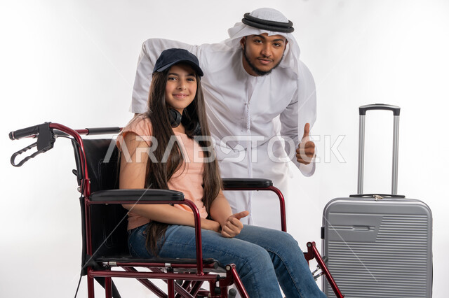 Psychological support and assistance to people with motor disabilities to move, Emirati society’s interest in people of determination, preparing to spend a tourist trip with people with special needs, portrait of a smiling, paralyzed Arab Gulf Emirati girl sitting in a wheelchair accompanied by her father, white background