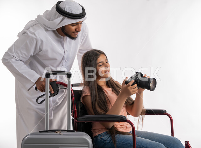 الاهتمام والعناية بذوي الاعاقة الحركية, تأمين اجهزة تساعد أصحاب الهمم على التنقل، رجل عربي خليجي إماراتي يرتدي الكندورة والغترة يستعد للسفر برحلة سياحية برفقة ابنته المقعدة, فتاة اماراتية تجلس على كرسي متحرك تمسك بيدها كاميرا احترافية عالية الدقة, خلفية بيضاء