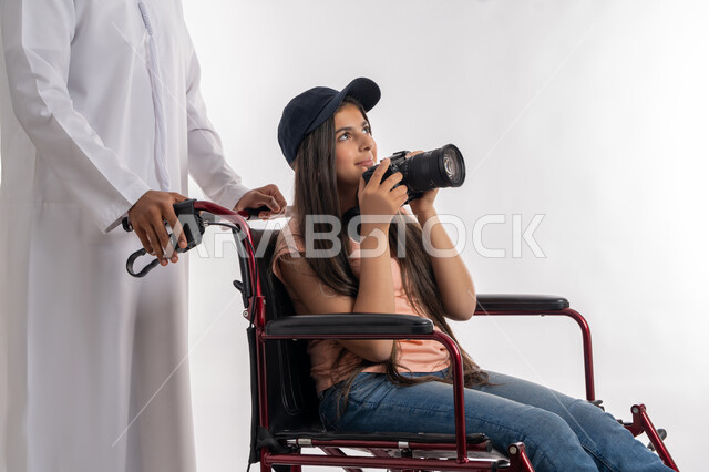 التقاط صور تذكارية باستخدام كاميرا احترافية عالية الدقة, تأمين اجهزة ومعدات تساعد ذوي الاحتياجات الخاصة على التنقل، بورتريه لرجل عربي خليجي إماراتي يرتدي الكندورة الاماراتية يقوم بمساعده ابنته المعاقة على الحركة باستخدام الكرسي المتحرك, خلفية بيضاء