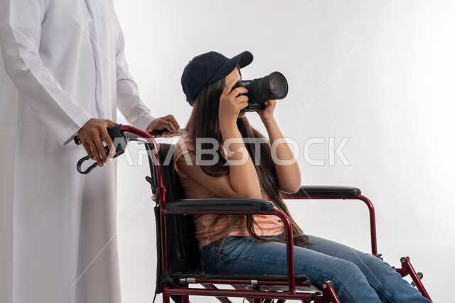 تأمين اجهزة تساعد ذوي الاحتياجات الخاصة على التنقل، التقاط صور تذكارية باستخدام كاميرا احترافية عالية الدقة, بورتريه لرجل عربي خليجي إماراتي يرتدي الكندورة الاماراتية يقوم بمساعده ابنته المعاقة على الحركة باستخدام الكرسي المتحرك, خلفية بيضاء