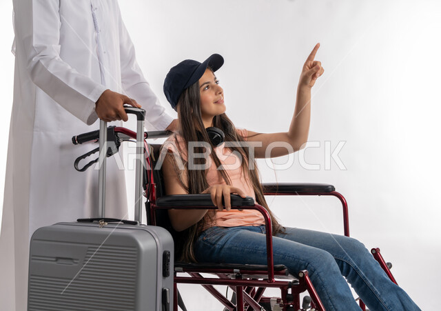 الاستمتاع بالرحلات السياحية برفقة الأبناء ذوي الاحتياجات الخاصة, بورتريه لرجل عربي خليجي إماراتي يرتدي الكندورة الاماراتية يمسك بيده حقيبة السفر مع ابنته المقعدة, الجلوس على كرسي متحرك والاشارة إلى شيء ما, تأمين معدات تساعد بالحركة والتنقل, خلفية بيضاء