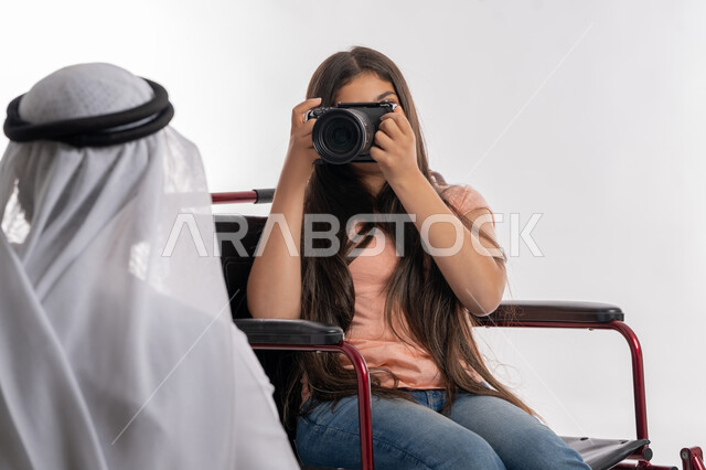 تنمية المواهب والهوايات المفضلة لذوي الهمم, التقاط  صور تذكارية عائلية, بورتريه لفتاة عربية خليجية إماراتية مقعدة تجلس على كرسي متحرك تمسك بيدها كاميرا احترافية عالية الدقة برفقة والدها, تقديم الحب والرعاية والاهتمام للأبناء المعاقين, خلفية بيضاء