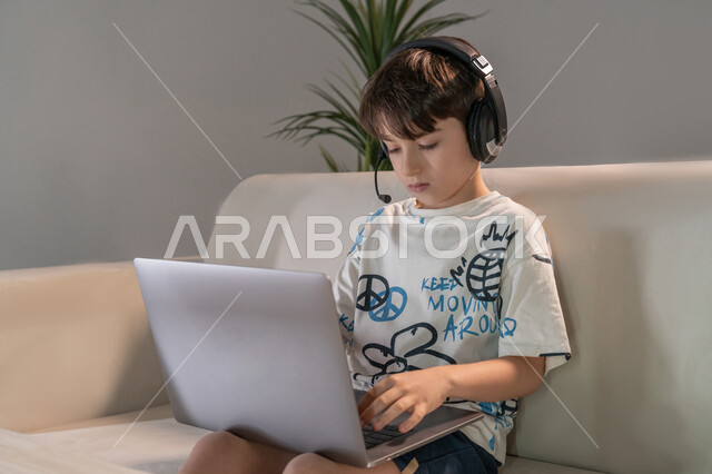 اللعب والتسلية بالألعاب الالكترونية المتطورة، قضاء اوقات ممتعة في العطلة، مشاهدة الافلام والمسلسلات الترفيهيه في المنزل، متابعة الدورات التعليمية لتنمية المهارات الفنية والفكرية، ولد عربي خليجي إماراتي يضع سماعة الهيدفون يستخدم الحاسب المحمول