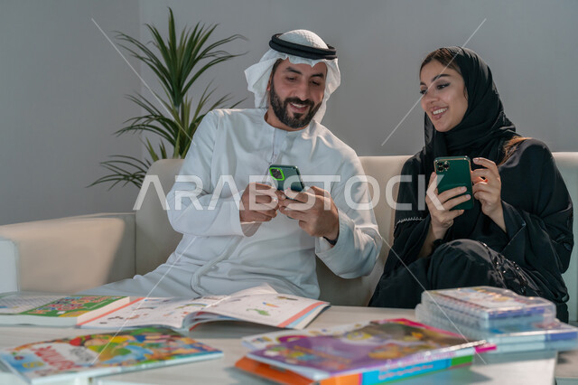 البحث عن دورات ومنصات تعليمية عبر الانترنت، زوجان عربيان خليجيان إماراتيان يقومان باستخدام الهاتف المحمول، طاولة منزلية مليئة بالكتب المدرسية، الاهتمام بتنمية وتطوير المهارات الفنية والعلمية للأبناء