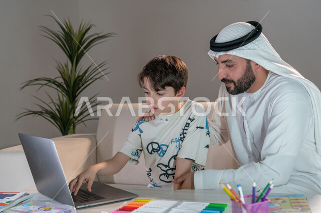 حل الواجبات المنزليه باستخدام الحاسب المحمول، أب عربي خليجي اماراتي يرتدي الكندورة والغترة يقوم بتعليم وتدريس ابنه، تنمية المهارات الثقافية والقدرات الفكرية للابناء، معلم خصوصي لمتابعة الدروس في المنزل، دمج التعليم مع التقنية والتكنولوجيا