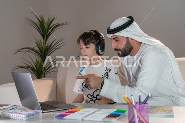 حل الواجبات المنزليه باستخدام الحاسب المحمول، أب عربي خليجي اماراتي يرتدي الكندورة والغترة يقوم بتعليم وتدريس ابنه، تنمية المهارات الثقافية والقدرات الفكرية للابناء، معلم خصوصي لمتابعة الدروس في المنزل، دمج التعليم مع التقنية والتكنولوجيا