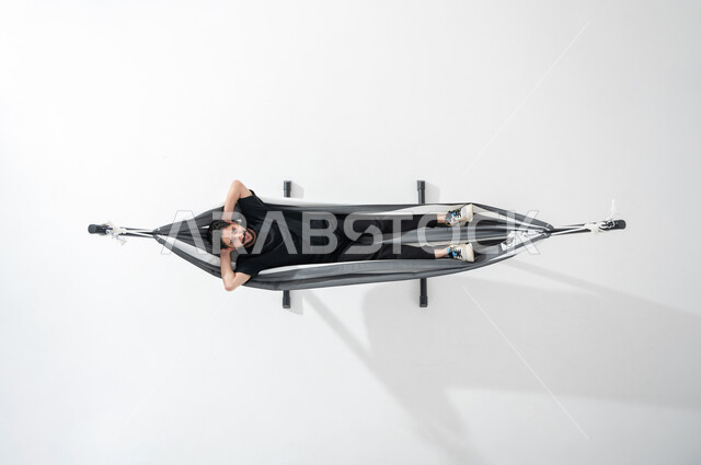 الاستمتاع في قضاء وقت الاستراحة، بورتريه عامودية علوية لشاب عربي خليجي سعودي مبتسم يستلقي على أرجوحة قماشية معلقة يرتدي ملابس سوداء وحذاء رياضي وينظر إلى الأعلى، وضع اليدين خلف الرأس, ايماءات تدل على الاسترخاء والهدوء، خلفية بيضاء