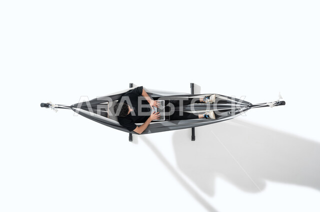 Taking advantage of technology and advanced techniques, an overhead portrait of a smiling young Arab Gulf Saudi man, lying on a hanging cloth hammock, wearing a casual outfit, looking at the laptop, working on the laptop, spending time with himself, completing tasks remotely, white background