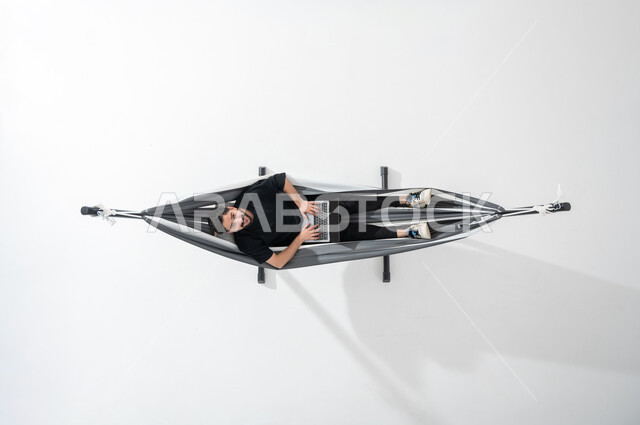 الاستفادة من التكنولوجيا والتقنيات المتطورة، بورتريه عامودية علوية لشاب عربي خليجي سعودي مبتسم يستلقي على أرجوحة قماشية معلقة يرتدي الزي الكاجوال ونظر إلى الأعلى، العمل على الحاسوب المحمول، قضاء الوقت مع الذات، انجاز المهام عن بعد، خلفية بيضاء