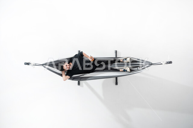 الاستمتاع واستغلال وقت الاستراحة، بورتريه عامودية علوية لشاب عربي خليجي سعودي مبتسم يستلقي على أرجوحة قماشية معلقة يرتدي الزي الكاجوال وينظر إلى الأعلى ويمسك بيده زجاجة عصير، القيام بأنشطة مختلفة، قضاء الوقت مع الذات، إيماءات تدل على الاسترخاء والفرح، خلفية بيضاء