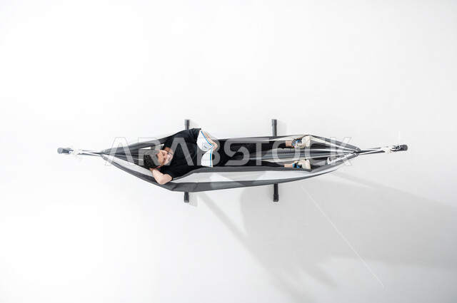 Relaxing and taking advantage of time by reading, taking a break, an overhead portrait of a Saudi Gulf Arab young man lying on a hanging cloth hammock, wearing black clothes and sports shoes, holding a book in his hand, placing his hand behind his head, learning and educating by reading cultural books, white background