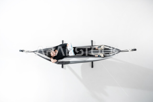 ممارسة هواية قراءة الكتب، استغلال وقت الاستراحة بالمطالعة, بورتريه علوي عامودي لشاب عربي خليجي سعودي مبتسم يرتدي الزي الكاجوال يجلس على أرجوحة قماشية معلقة ويمسك بيده مجلة ثقافية, النظر إلى الأعلى ووضع اليد خلف الرأس, قضاء الوقت بعيداً عن الأجهزة الذكية