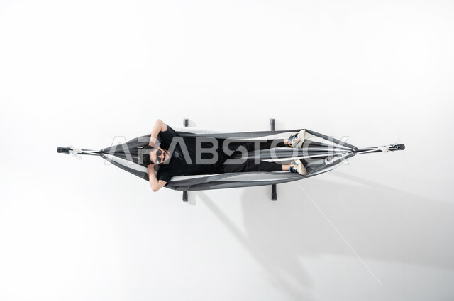 الاسترخاء والاستمتاع بأوقات الاستراحة، بورتريه لشاب عربي خليجي سعودي مبتسم يرتدي الزي الكاجوال ويستلقي على أرجوحة قماشية معلقة يضع النضارات الشمسية، الشعور بالراحة والهدوء, قضاء الوقت بعيداً عن الآخرين، النظر إلى الأعلى وفتح اليدين والقدمين، خلفية بيضاء