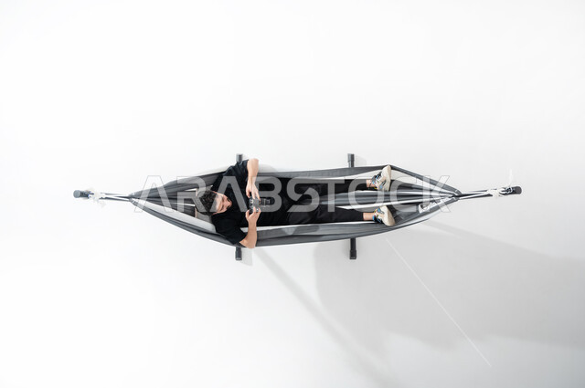 ممارسة الهوايات في أوقات الاستراحة, بورتريه عامودي علوي لشاب عربي خليجي سعودي يرتدي الثوب التقليدي ويستلقي على أرجوحة قماشية معلقة يمسك بيده آلة التصوير الرقمية, التقاط الصور الفوتوغرفية من خلال الكاميرا, تقريب الأجسام البعيدة, قضاء الوقت مع الذات