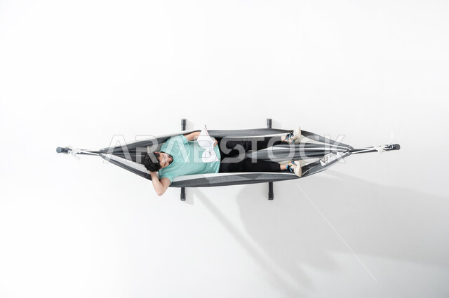 Taking advantage of time and practicing the hobby of reading books, taking advantage of break time by reading, a vertical overhead portrait of a smiling Saudi Gulf Arab young man wearing a casual outfit sitting on a hanging cloth hammock and holding a cultural magazine in his hand, looking at the magazine and placing the hand behind the head, spending time away from smart devices