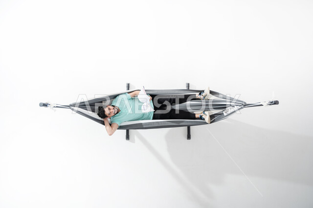 Practicing the hobby of reading books, using the break time to read, a vertical overhead portrait of a smiling Saudi Gulf Arab young man wearing a casual outfit sitting on a hanging cloth hammock and holding a cultural magazine in his hand, looking up and placing the hand behind the head, spending time away from smart devices