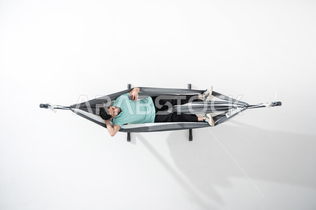 التسلية والمتعة خلال فترة الاستراحة, بورتريه عامودي علوي لشاب عربي خليجي سعودي مبتسم يرتدي الزي الكاجوال يستلقي على أرجوحة قماشية معلقة ويضع يده خلف رأسه، الامساك بالهاتف الذكي, استخدام الأجهزة الحديثة والمتطورة,  قضاء الوقت مع الذات، النظر إلى الموبايل، خلفية بيضاء