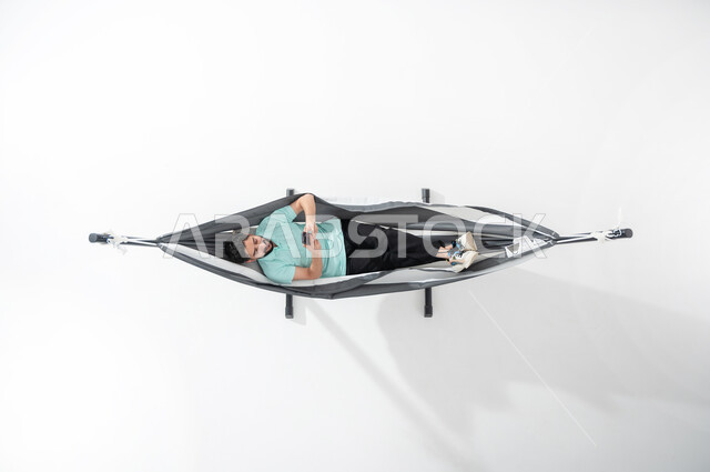 Relaxation and calm during the break, a vertical portrait of a smiling Saudi Gulf Arab young man wearing a casual outfit, lying on a hanging cloth hammock and using a smartphone, using modern and advanced devices, the pleasure of spending time with oneself, looking at the mobile phone, a white background