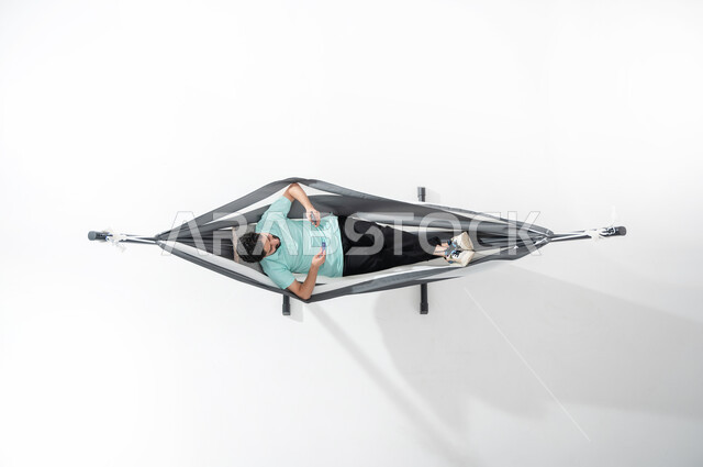Taking advantage of technology and modern techniques, a vertical overhead portrait of a Saudi Gulf Arab young man wearing a casual outfit sitting on a hanging cloth hammock and making electronic payments using a mobile phone, using an ATM card, ease of paying via a bank account, white background