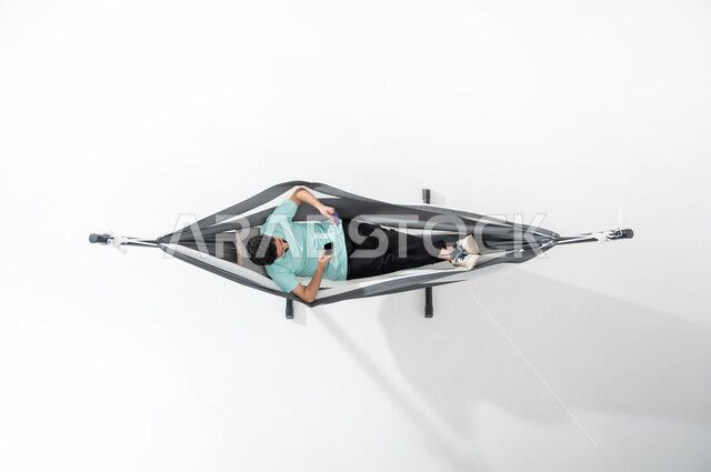 التكنولوجيا المتطورة والتقنيات الحديثة, بورتريه علوي عامودي لشاب عربي خليجي سعودي  يرتدي الزي الكاجوال يجلس على أرجوحة قماشية معلقة ويقوم بالدفع الالكتروني بواسطة الهاتف المحمول, استخدام بطاقة الصراف, سهولة الدفع عبر الحساب البنكي, خلفية بيضاء