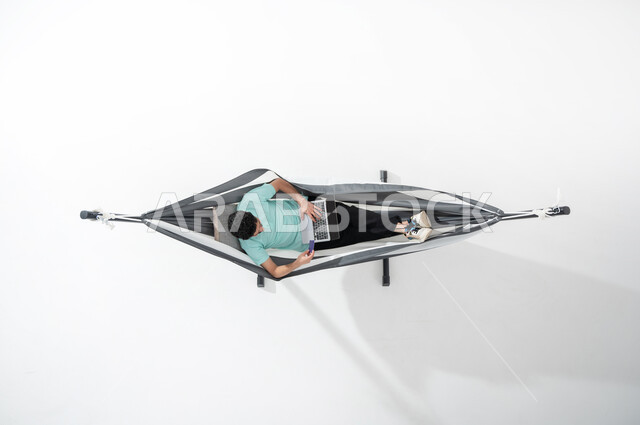 الاستفادة من التكنولوجيا المتطورة والتقنيات الحديثة, بورتريه علوي عامودي لشاب عربي خليجي سعودي  يرتدي الزي الكاجوال يجلس على أرجوحة قماشية معلقة ويقوم بالدفع الالكتروني بواسطة الحاسوب المحمول, استخدام بطاقة الصراف, سهولة الدفع عبر الحساب البنكي, خلفية بيضاء