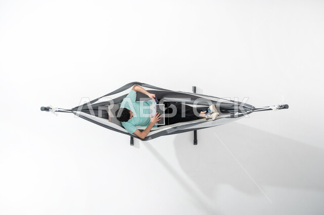 استخدام التكنولوجيا المتطورة والتقنيات الحديثة, بورتريه علوي عامودي لشاب عربي خليجي سعودي  يرتدي الزي الكاجوال يجلس على أرجوحة قماشية معلقة ويقوم بالدفع الالكتروني بواسطة الحاسوب المحمول, استخدام بطاقة الصراف, سهولة الدفع عبر الحساب البنكي, خلفية بيضاء