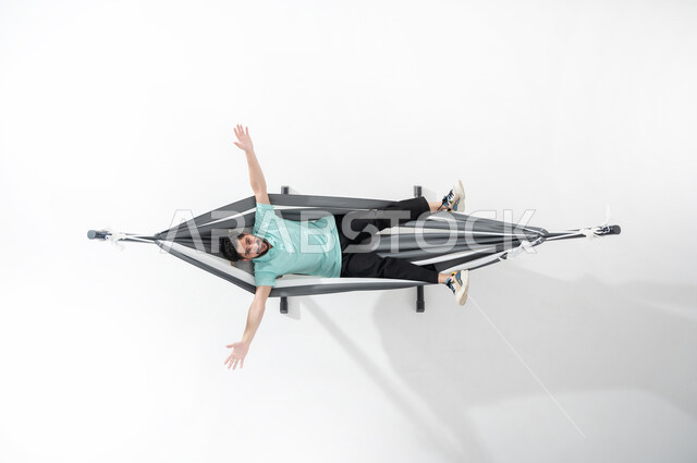 الاستمتاع بأوقات الاستراحة، إيماءات تدل على السعادة والبهجة، بورتريه لشاب عربي خليجي سعودي مبتسم يرتدي الزي الكاجوال ويستلقي على أرجوحة قماشية معلقة، الشعور بالراحة والهدوء, قضاء الوقت بعيداً عن الآخرين، النظر إلى الأعلى وفتح اليدين والقدمين، خلفية بيضاء