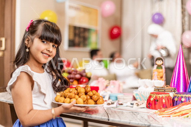 طفلة سعودية خليجية سعيدة ترتدي لباس العيد الانيق ، تشارك الاجواء الاحتفالية الخاصة بعيد الفطر المبارك ، سفرة مليئة بأصناف الحلويات ، الاحتفال بعيد الفطر السعيد ، عيد الفطر المبارك  ،  تقديم العيديات و الحلوى ، القرقيعان