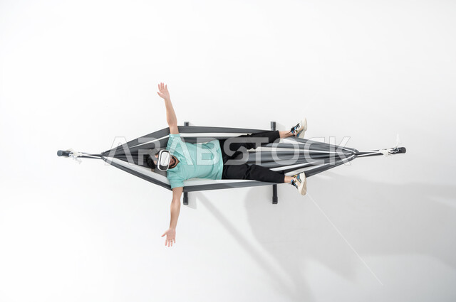 Using technology for entertainment and amusement, enjoying break times, an overhead portrait of a smiling Saudi Gulf Arab young man wearing casual clothes and lying on a hanging cloth hammock wearing virtual reality glasses, hand gestures indicating rest and relaxation, white background