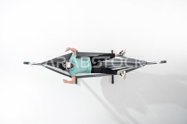 الاستمتاع بأوقات الاستراحة, بورتريه عامودي علوي لشاب عربي خليجي سعودي مبتسم يرتدي الزي الكاجوال ويستلقي على أرجوحة قماشية معلقة يضع نظارات الواقع الافتراضي، ايماءات اليدين تدل على الإعجاب والموافقة، استخدام التكنولوجيا في الترفيه والتسلية، خلفية بيضاء