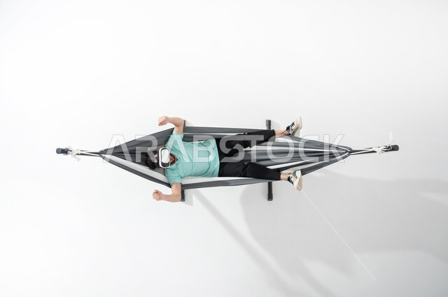 Relaxing and enjoying break times, a vertical portrait of a smiling Saudi Gulf Arab young man wearing casual clothes and lying on a hanging cloth hammock wearing virtual reality glasses, hand gestures indicating enthusiasm and joy, using technology for entertainment and amusement, white background
