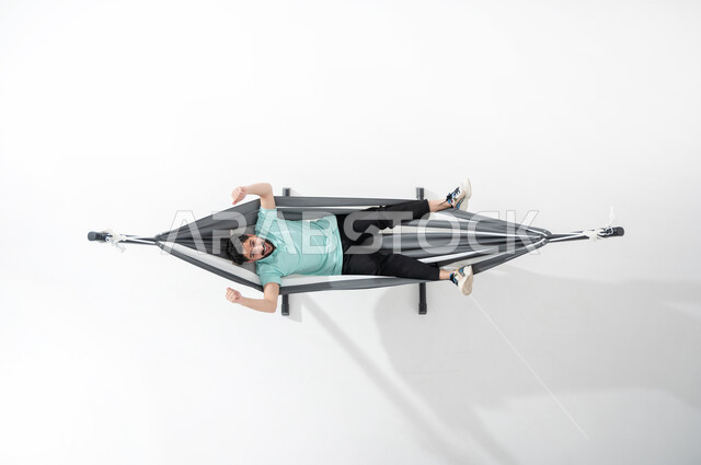 الاستمتاع بأوقات الاستراحة, بورتريه عامودي علوي لشاب عربي خليجي سعودي مبتسم يرتدي الزي الكاجوال ويستلقي على أرجوحة قماشية معلقة، ايماءات اليدين تدل على الحماس والبهجة، النظر إلى الأعلى، قضاء الوقت مع الذات، خلفية بيضاء