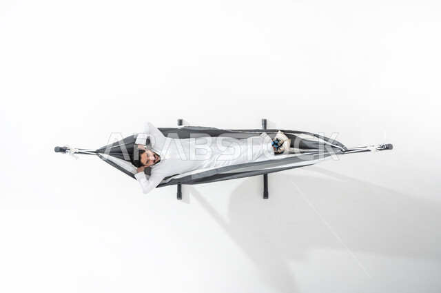 الاسترخاء والاستمتاع بأوقات الاستراحة, بورتريه عامودي علوي لشاب عربي خليجي سعودي مبتسم يرتدي الثوب التقليدي ويستلقي على أرجوحة قماشية معلقة، ويضع يديه خلف رأسه، رؤية مستقبلية سعودية, ايماءات تدل على الراحة والسرور, النظر إلى الأعلى