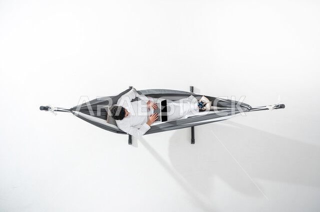 Using advanced technology and modern techniques, a vertical portrait of a smiling Saudi Gulf Arab young man wearing a traditional dress, sitting on a hanging cloth hammock and using a laptop, doing work remotely, white background