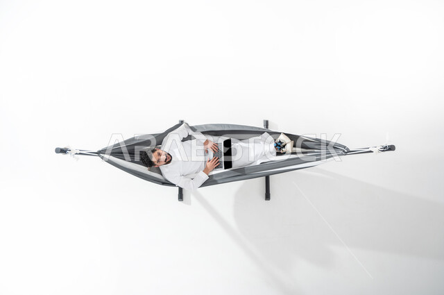 الاستفادة من التكنولوجيا المتطورة والتقنيات الحديثة, بورتريه علوي عامودي لشاب عربي خليجي سعودي مبتسم يرتدي الثوب التقليدي يجلس على أرجوحة قماشية معلقة ويقوم بالدفع الالكتروني بواسطة الحاسوب المحمول, استخدام بطاقة الصراف, سهولة الدفع عبر الحساب البنكي, خلفية بيضاء